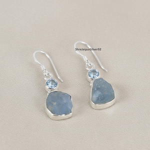 Puede incluir: Un par de pendientes colgantes de plata. Cada pendiente presenta una piedra azul claro de forma irregular engastada en un bisel plateado, con una piedra azul redonda más pequeña encima. Los pendientes tienen un diseño de gancho simple.
