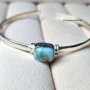 Könnte beinhalten: Ein silbernes Armreif-Armband mit einem quadratischen, hellblauen Edelstein in der Mitte. Der Edelstein ist in einer silbernen Lünette gefasst und wird von dekorativen silbernen Akzenten flankiert. Das Armband hat eine glatte, polierte Oberfläche.