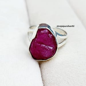 May include: A silver ring with a large, raw-cut ruby gemstone. The ruby is a deep magenta hue, set in a polished silver band with a split shank design. The ring is presented on a white background.