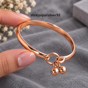 May include: A rose gold-toned bangle bracelet featuring a smooth, polished finish and a circular charm connector. Two small, spherical rose gold-toned beads hang from the connector. The text shrejaipursilver92 appears in the upper center area.