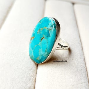 May include: A large oval turquoise ring with a silver band. The turquoise stone is a vibrant blue-green colour with brown and gold veining. The ring is set on a white surface.