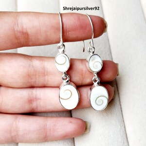 May include: A pair of drop earrings featuring two oval-shaped white Shiva eye shell cabochons set in polished silver. Each earring has a hook-style wire and displays a natural spiral pattern on the shell surface. The text Shrejaipursilver92 is visible.