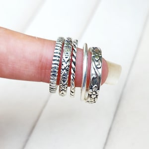 May include: A collection of silver rings with various designs. The rings include textured, braided, and floral patterns. The rings are displayed on a finger, showcasing their different styles and textures.