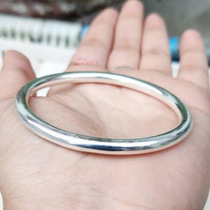 Può includere: Un braccialetto a cerchio color argento con un design liscio e arrotondato. Il braccialetto è circolare e sembra essere fatto di metallo. Il braccialetto è tenuto in una mano, mostrando la sua superficie lucida e il suo stile semplice ed elegante.