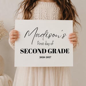 Op de afbeelding: Een rechthoekig wit bord met zwarte tekst die luidt: Madison's First day of SECOND GRADE 2026-2027. Het bord bevat een combinatie van elegant cursief schrift voor de naam en een vetgedrukte, duidelijke typografie voor het leerjaar.