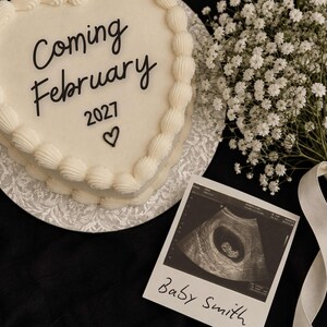 Puede incluir: Un pastel en forma de corazón con glaseado blanco y bordes decorados, que presenta el texto negro "Coming February 2027" y un pequeño icono de corazón. Al lado hay una ecografía en blanco y negro con el texto escrito a mano "Baby Smith".
