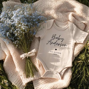 Pode incluir: Um body de bebé bege com o texto "Baby Anderson" e "Due October 2023" é exibido sobre uma manta de malha creme. Um buquê de flores azuis amarrado com uma fita branca e pequenas flores brancas também estão na imagem.