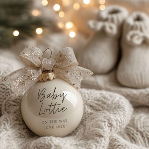 Puede incluir: Una bola de Navidad color crema con el texto "Baby Lottie ON THE WAY JUNE 202X" en escritura negra. El adorno tiene una parte superior dorada y está adornado con un lazo transparente y brillante. Dos pares de patucos de bebé están en el fondo.