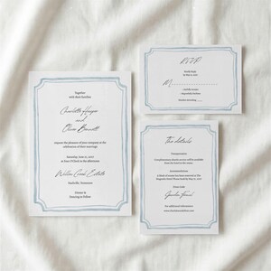 Op de afbeelding: Een set van drie witte trouwkaarten met een lichtblauwe, handgeschilderde rand. De hoofduitnodiging, RSVP-kaart en detailkaart bevatten zwarte gedrukte tekst met namen, evenementinformatie en een websiteadres voor de gasten.