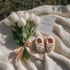 May include: A display featuring white tulips, a card reading "Baby Valentina", and two pairs of knitted baby booties with pink bows, set on a cream-coloured blanket.