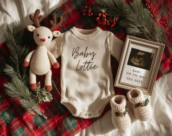 May include: A cream-colored baby onesie with the text "Baby Lottie" is displayed with a framed ultrasound image, a crocheted reindeer, and matching booties. The items are arranged on a red and green plaid blanket with pine and berry accents.