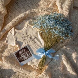 May include: A cream-coloured ribbed baby bodysuit featuring the black printed text Baby Miller JUNE 2026, accompanied by a bouquet of light blue baby's breath flowers tied with a matching blue ribbon and a square ultrasound scan photograph.