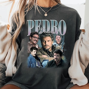 May include: Dark gray sweatshirt featuring a collage of images of Pedro Pascal. The word "PEDRO" is printed in large, stylized letters above the images, with "Pascal" below. The images show Pascal in various poses and expressions.