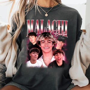 May include: Dark grey sweatshirt with a graphic of a young man, the name "Malachi Barton" in a vintage style. The design includes multiple images of the person. The sweatshirt has a crew neck and long sleeves.