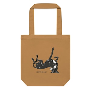 May include: A tan tote bag featuring a black and white graphic illustration of a dog lying on its back with its legs in the air. Below the dog, the text "I'D RATHER NOT" is printed in a simple black font. The bag has two matching tan handles.