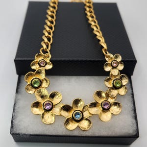 May include: A gold-tone chain necklace featuring a series of five-petaled flower charms. Each flower has a textured gold finish and a central round gemstone in varying colors including light blue, pink, and green, set within a black border.