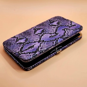 May include: A purple and black snakeskin-patterned clutch wallet. The wallet is rectangular with a hinged opening and a silver-colored clasp. The interior appears to be black. The wallet is open, revealing the interior.