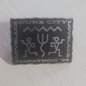 May include: A silver pin with a black background. The pin features the text "Duke City Lovelace Marathon '92" and a stylized image of two figures with a trident.