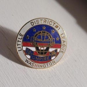 May include: A round pin with a gold border featuring the text "LITTLE LEAGUE DISTRICT #1" and "UMPIRE WASHINGTON." The pin has a blue background with gold stars and a red and white stripe design. A baseball umpire mask is in the center.