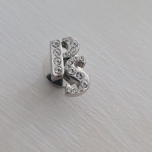 Könnte beinhalten: Ein silberner Anhänger mit den Buchstaben "RS", besetzt mit klaren Strasssteinen.