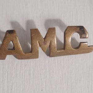 Puede incluir: Un emblema metálico envejecido con las letras "AMC" en una fuente estilizada. Las letras están unidas y tienen un acabado similar al bronce, mostrando signos de desgaste. Podría ser una insignia de coche o un artículo vintage similar.