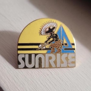 Puede incluir: Un pin de esmalte amarillo, dorado y azul con la palabra "SUNRISE" en letras plateadas. El pin presenta una figura estilizada con un tocado y un diseño de triángulo azul.