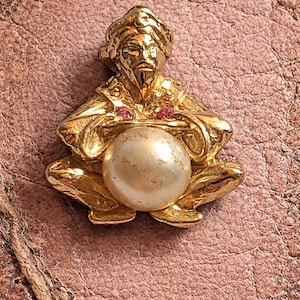 Peut inclure: Figurine dorée représentant une figure assise tenant une grosse perle. La figure a une barbe et porte un chapeau. Deux petites pierres précieuses rouges sont visibles. La figurine est sur une surface brune texturée.