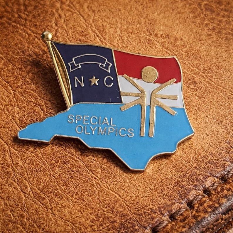 May include: A gold-toned pin in the shape of the state of North Carolina. The pin features a flag design with the letters "N C" and the words "Special Olympics" in blue. The flag has red, white, and blue sections, and a gold-colored figure.