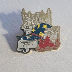 May include: A gold-colored pin with a white castle, a blue hat with stars, and a red dragon. The pin has the text "Amaranth Makes Magic" on a white banner.