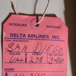 Pode incluir: Uma etiqueta de bagagem rosa vintage da Delta Airlines com detalhes manuscritos. A etiqueta indica "SAN DIEGO" como destino final, com informações de voo e uma indicação de peso. Uma corda está presa.
