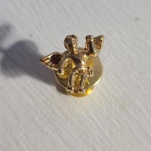 May include: A gold-coloured angel pin with outstretched arms and small wings. The pin is a detailed miniature sculpture of a cherub, mounted on a circular base. The pin is likely made of metal.