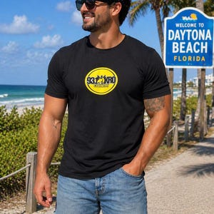 May include: Black t-shirt with a yellow circular logo that reads "93.1 KRO Country". The person is wearing blue jean shorts and sunglasses. The background includes a beach, palm trees, and a sign that says "Welcome to Daytona Beach Florida".