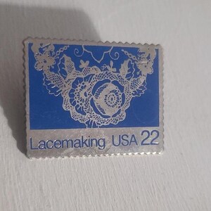May include: A silver-coloured pin with a blue background featuring a detailed lace design. The pin is rectangular with a scalloped edge, and the text "Lacemaking USA 22" is printed at the bottom.