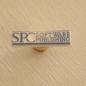 Pode incluir: Um alfinete retangular com o texto "SPC SOFTWARE PUBLISHING" em letras azuis. O alfinete tem um fundo branco com uma borda azul e um suporte de alfinete dourado.