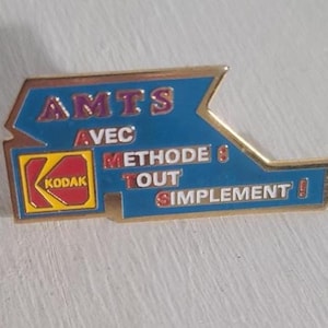 Könnte beinhalten: Ein Vintage-Kodak-Anstecknadel mit blauem und goldenem Design. Die Anstecknadel zeigt den Text "AMTS AVEC METHODE 8 TOUT SIMPLEMENT!" und das Kodak-Logo in einem roten und gelben Quadrat.