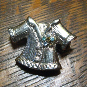 May include: A silver-toned brooch shaped like a jacket with a decorative rhinestone design in the center.