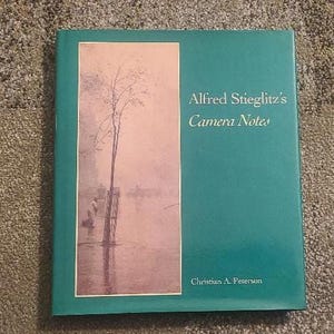 May include: A teal book titled "Alfred Stieglitz's Camera Notes" with a landscape image on the left. The image shows a tree in water with a pink and white background. The author's name, Christian A. Peterson, is at the bottom.