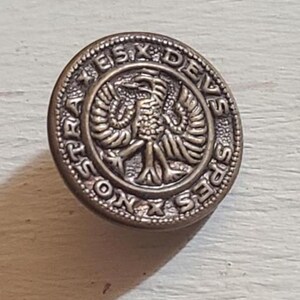Könnte beinhalten: Ein runder, antiker Messingknopf mit erhabenem Text und einem zentralen Emblem. Der Text am Rand lautet "SPES NOSTRA DEUS". Das Emblem in der Mitte scheint ein stilisierter Vogel mit ausgebreiteten Flügeln zu sein.