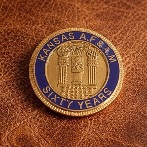 Puede incluir: Una moneda dorada con texto azul y blanco que dice "KANSAS A.F.&A.M. SIXTY YEARS". La moneda presenta un diseño central con pilares y un símbolo. La moneda está sobre un fondo de cuero marrón.