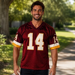 May include: A maroon football jersey with the number 14 in white, accented with gold and white on the sleeves. The jersey has a V-neck design and is made of a breathable material.
