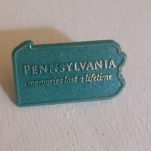 May include: Teal-coloured magnet in the shape of Pennsylvania, featuring the words "PENNSYLVANIA" and "memories last a lifetime" in gold. The magnet has a textured surface and a raised border.