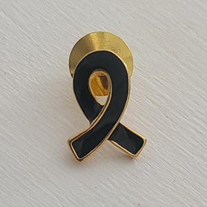 May include: Black ribbon pin with a gold backing. The ribbon is a symbol of mourning and remembrance.