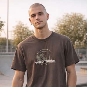 May include: A brown t-shirt with the text "volcomstone" in white, featuring a circular design. The shirt is worn by a person with short hair and earrings, standing outdoors.