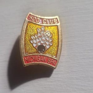 May include: A gold-colored pin with a bowling theme. The pin features a bowling ball and pins in the center, with the text "500 CLUB" at the top and "MICHIGAN BPA" at the bottom in red lettering. The background is textured gold.