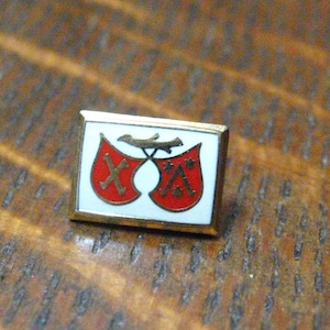 Op de afbeelding: Een gouden speld met een witte achtergrond en twee rode schilden. Elk schild heeft een gestileerde 'X' en 'A' in het midden.