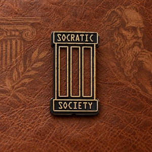 Könnte beinhalten: Eine rechteckige Anstecknadel aus Metall mit schwarz-goldener Oberfläche und drei vertikalen Aussparungen in der Mitte. Das Wort SOCRATIC ist oben in Gold geprägt, das Wort SOCIETY unten in Gold, umrahmt von einer goldenen Umrandung.