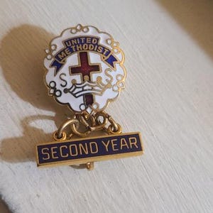 May include: A gold-toned United Methodist pin with a white and blue emblem featuring a cross and crown. The pin has a banner that reads "SECOND YEAR" in gold lettering on a navy blue background.