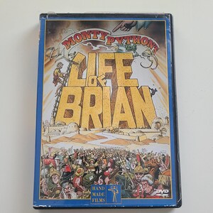 Puede incluir: Una caja de DVD para la película de Monty Python "La Vida de Brian". La caja presenta una imagen de dibujos animados del título de la película en letras grandes y amarillas sobre un fondo de desierto. La caja es azul con un borde blanco y las palabras "Hand Made Films" están impresas en la parte inferior.