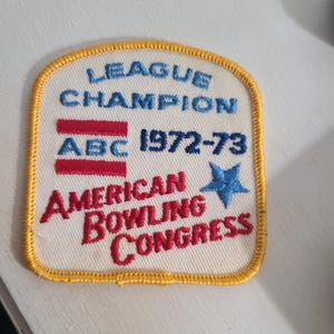 May include: A vintage, rectangular patch with a cream-colored background and a yellow stitched border. The patch features the text "LEAGUE CHAMPION ABC 1972-73 AMERICAN BOWLING CONGRESS" in blue and red lettering, with a blue star.