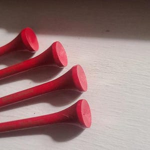 Może przedstawiać: Cztery czerwone kołki golfowe ułożone są po przekątnej na białej powierzchni. Każdy kołek ma długą, smukłą n&oacute;żkę i płaską, okrągłą gł&oacute;wkę. Kołki wykonane są z drewna lub podobnego materiału.